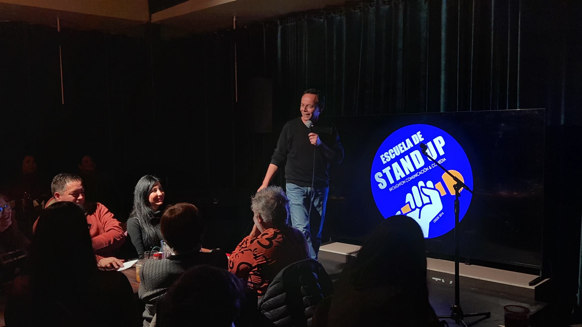 Raúl “Gato Viejo” Broughton en el escenario frente al público, con el logo de la Escuela de Stand Up en pantalla